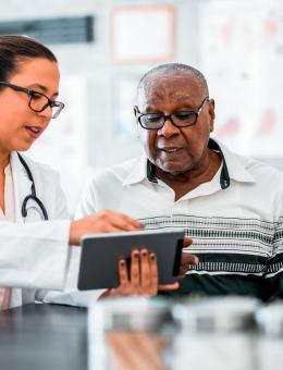 A man and a woman look at a tablet, the woman is a medical professional and the man is of retirement age