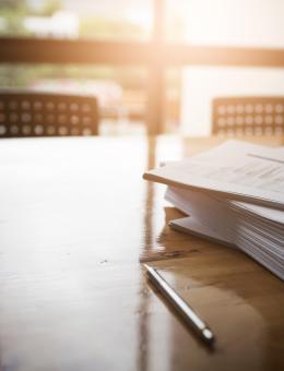 Papers on a desk in front of a window with sun shining through. Glasses on top of the papers and pens on the desk