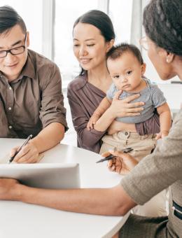 A woman is presenting a document to a man who is signing it, while another woman holds a baby.