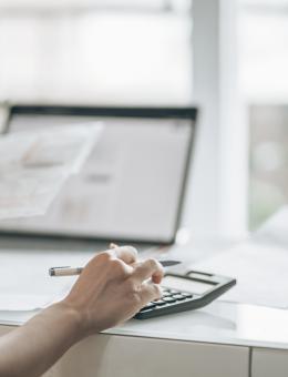 Woman seen from behind using a calculator, looking at a document with a computer open